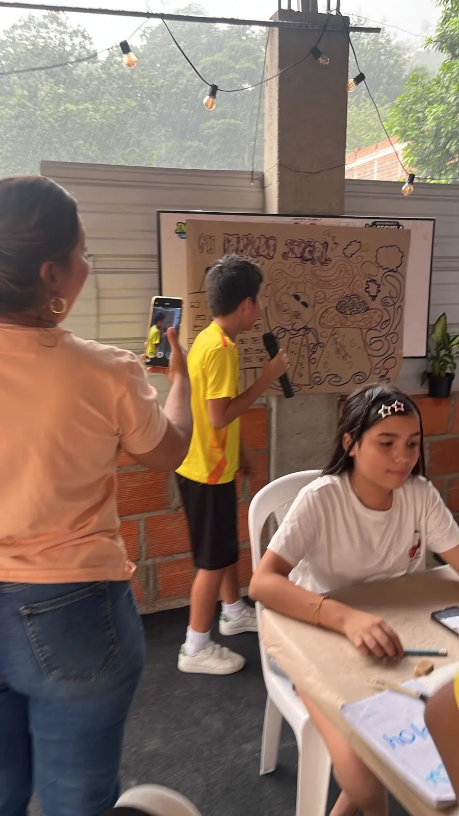 Un estudiante expone su mural «Mi mundo ideal» con micrófono mientras llueve